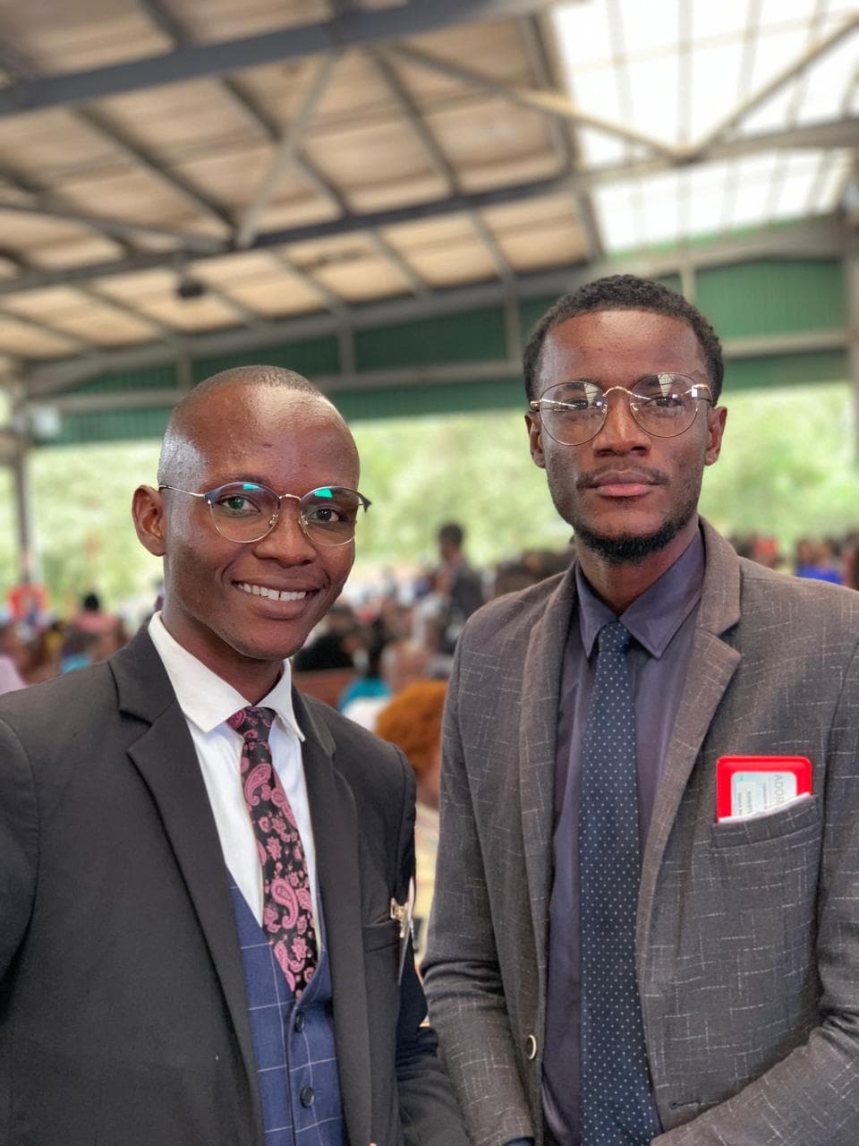 Dois homens negros jovens vestindo ternos e óculos, sorrindo em um grande pavilhão coberto.